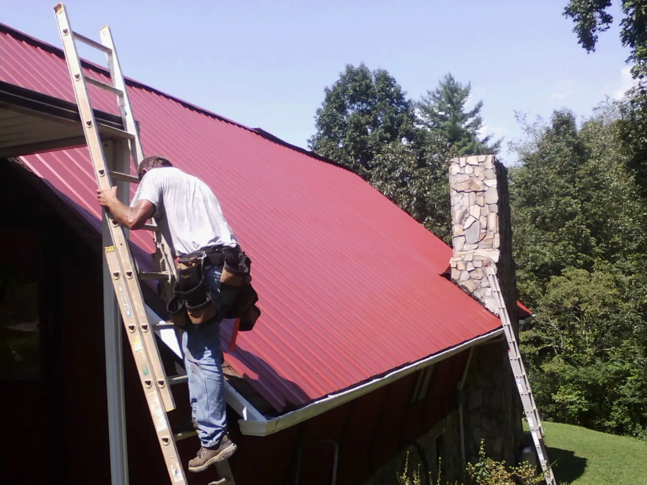 Roofer on a ladder beside a red metal roof and stone chimney — Soffit & Fascia Repair by Roofing Brothers in Streamwood
